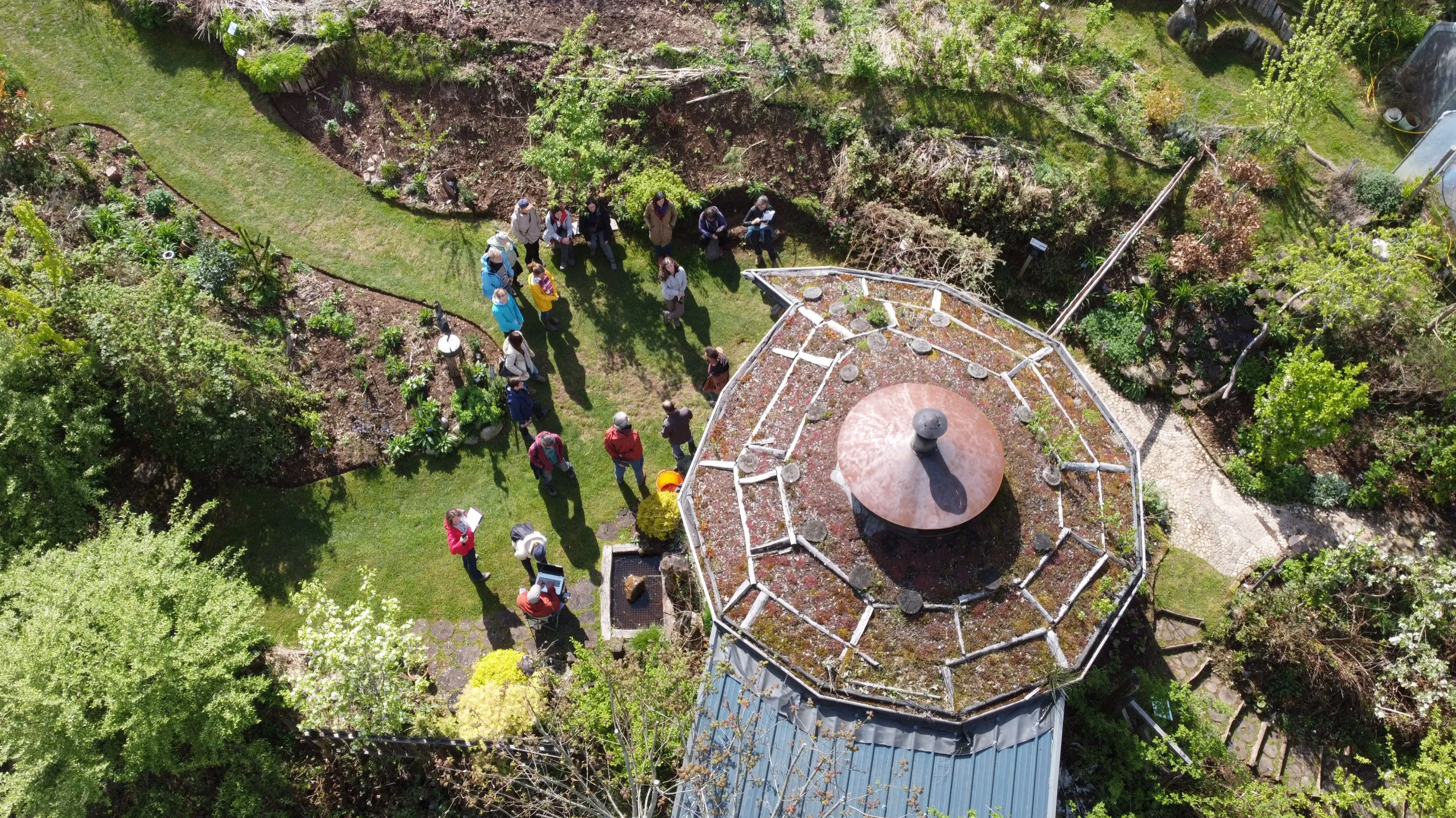 Vue aérienne d'un jardin-forêt mature : le groupe du cursus en visite près d'un kiosque au toit végétalisé